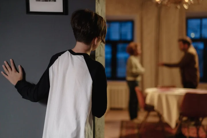 Dziecko podgląda zza framugi, w tle dwoje dorosłych kłócą się przy stole.