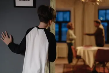 Dziecko podgląda zza framugi, w tle dwoje dorosłych kłócą się przy stole.