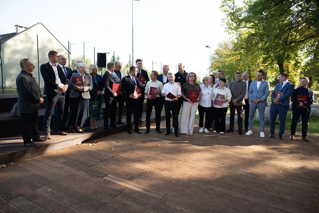 Grupa osób stoi na drewnianym podium na zewnątrz, większość trzyma bordowe dyplomy lub okładki, podczas uroczystości wręczenia wyróżnień w otoczeniu zieleni i o