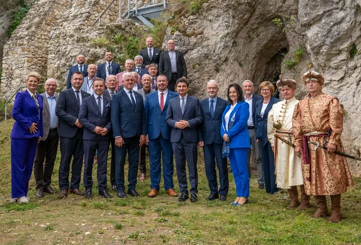 Grupa osób, głównie w garniturach, pozuje do pamiątkowego zdjęcia na tle skał i ruin, a obok stoją dwaj mężczyźni w historycznych strojach.
