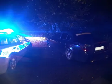 Policyjny radiowóz z oznaczeniem P028 stoi nocą przy czarnym samochodzie osobowym z otwartą pokrywą bagażnika na drodze w pobliżu zadrzewionego terenu.