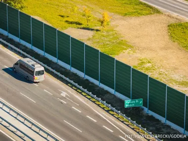 Bus jedzie po wielopasmowej drodze obok zielonego ekranu dźwiękochłonnego, przy którym stoi znak kierunkowy na Piekary Śl. i Będzin.